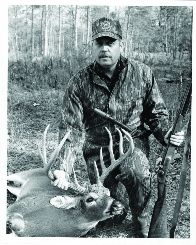 Isang itim at puting larawan ng isang lalaki, isang usa at isang muzzle loading rifle