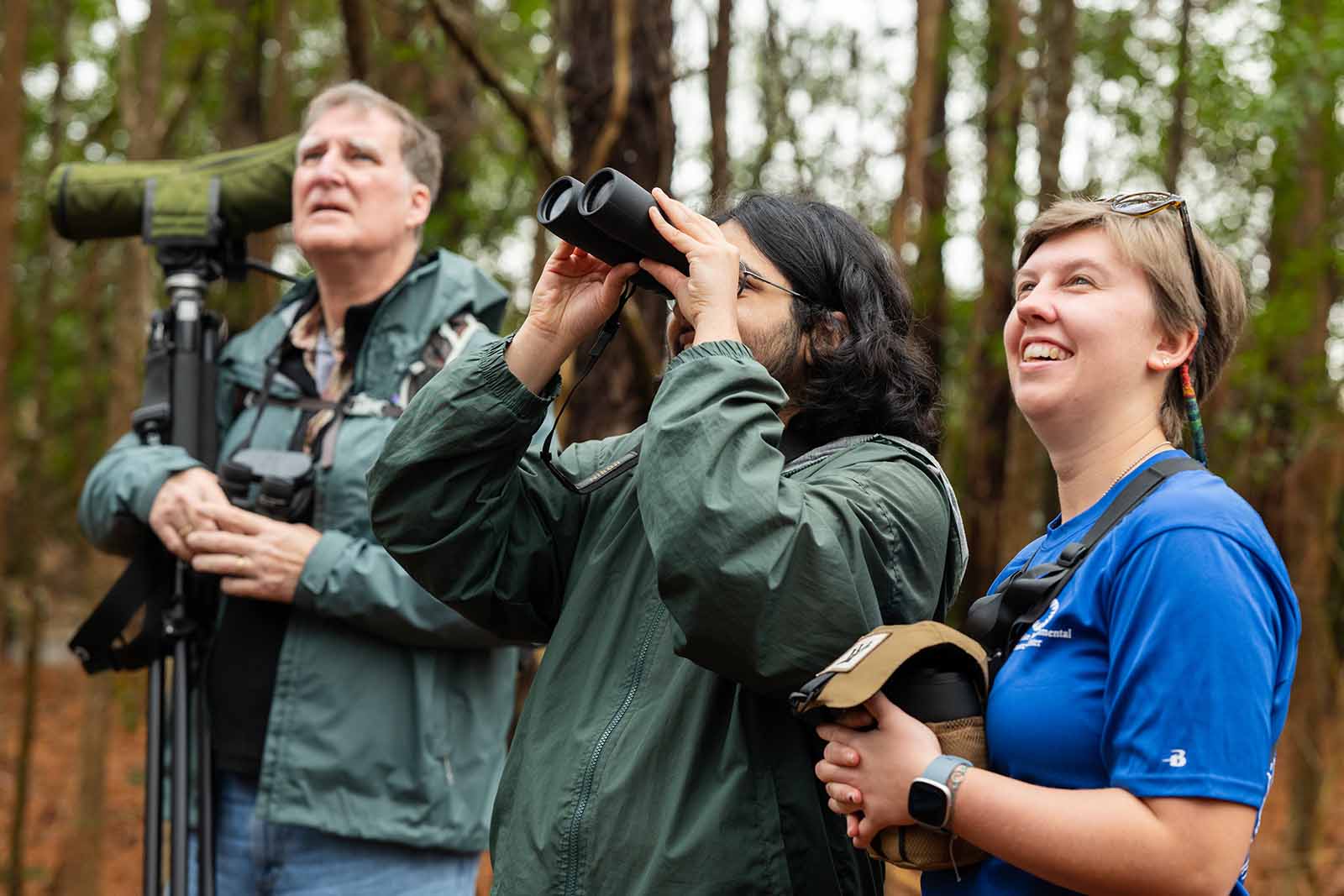 Dalawang tao na nakasuot ng dark green na jacket na nakatingin sa itaas, ang isa ay gumagamit ng isang pares ng binocular