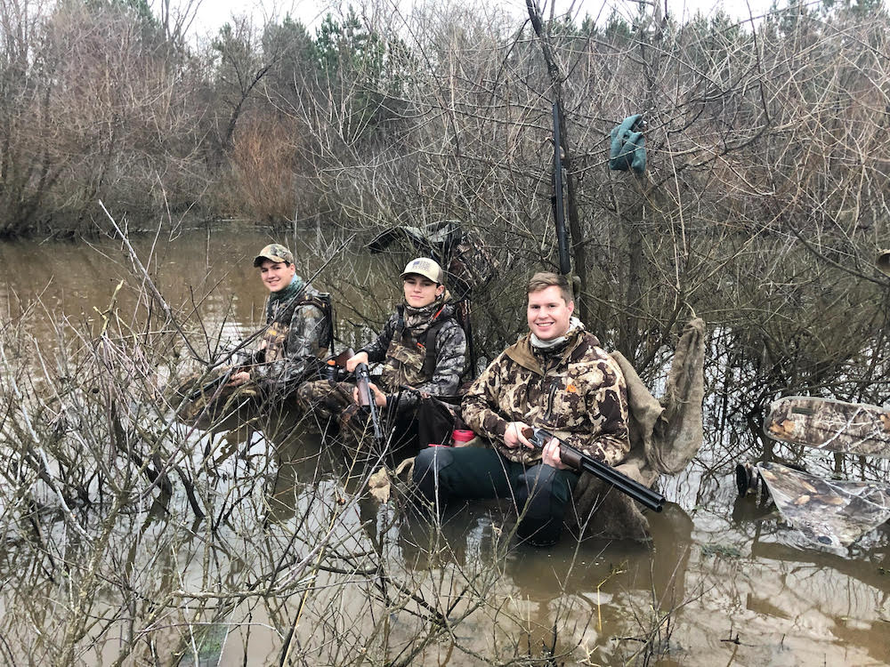 Isang imahe ng isang grupo ng mga lalaki na nakaupo sa isang troso sa tubig, wala silang nakitang pato sa pagkakataong ito