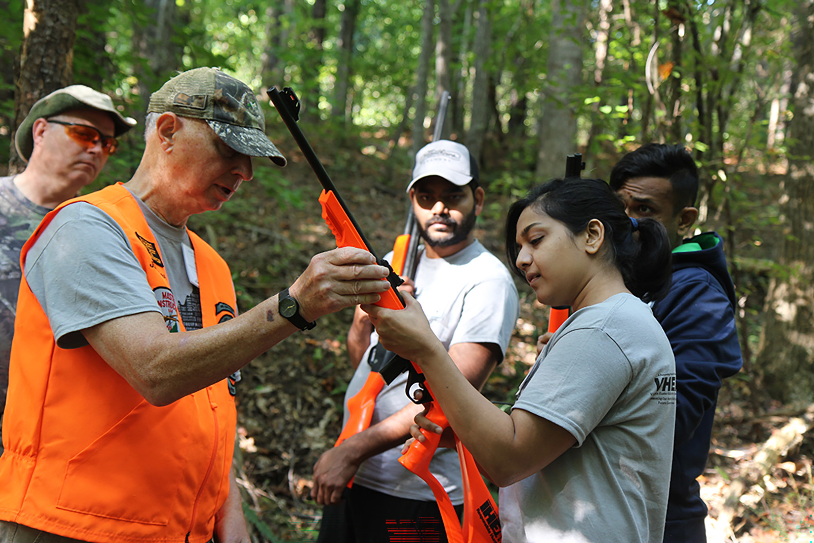 Isang larawan ng isang lalaking nagpapaliwanag kung paano humawak ng shotgun sa isang babae habang nakatingin ang isang grupo.