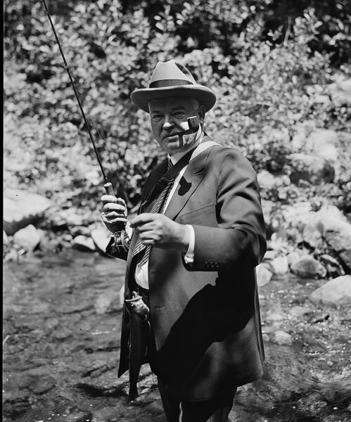 Isang itim-at-puting larawan ni Pangulong Herbert Hoover, nakasuot ng suit at may tubo sa kanyang bibig, nakatayo sa isang ilog na may hawak na fly rod at isang trout. 