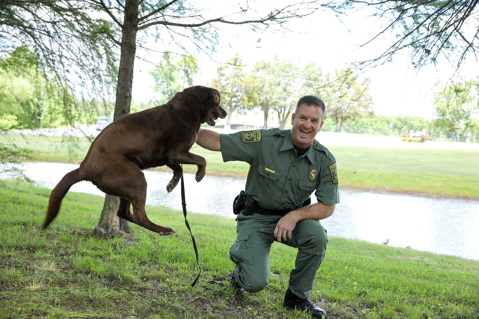 Isang larawan ng isang brown Labrador Retriever na tuwang-tuwa na tumatalon sa tabi ng nakaluhod na Conservation Police Officer. 