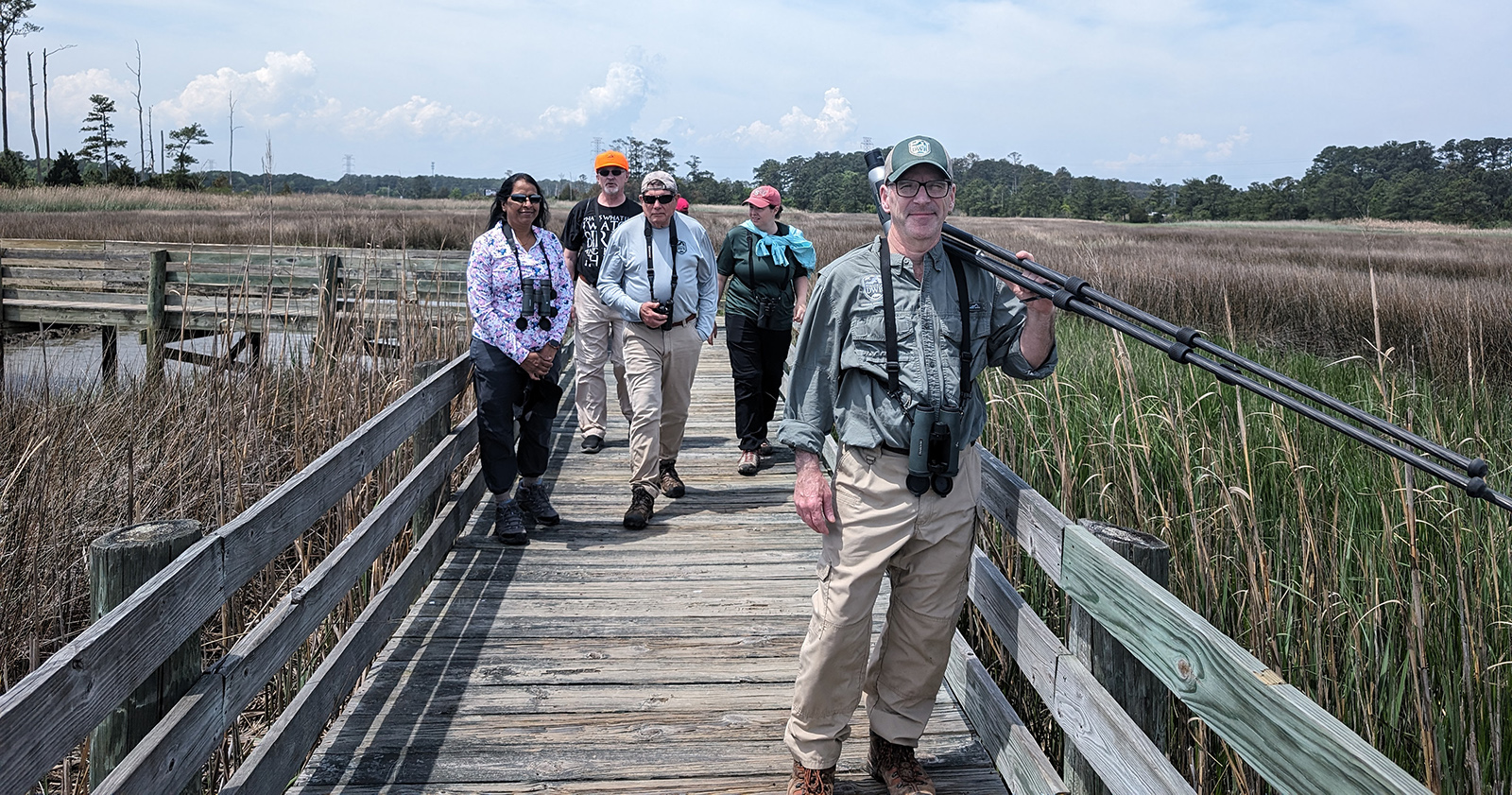 Isang larawan ng limang tao na may binocular na naglalakad sa isang kahoy na boardwalk sa isang wetland.