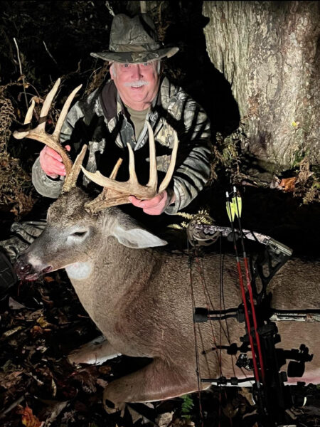 Isang larawan ng isang lalaking naka-camouflage na may busog na hawak ang mga sungay ng isang malaki, inani na buck.