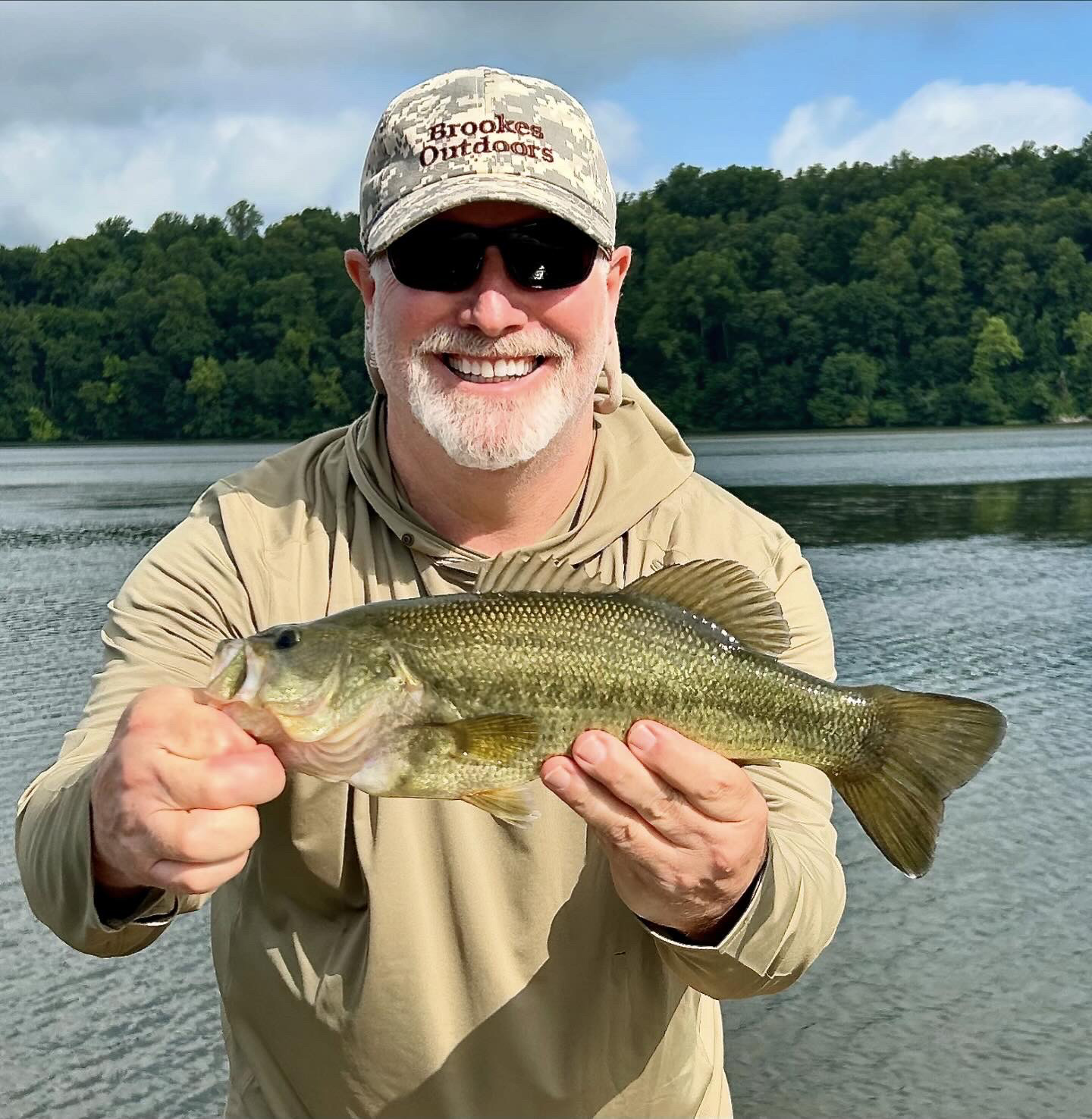Isang larawan ng nakangiting lalaki na may hawak na largemouth bass na may ilog sa background.
