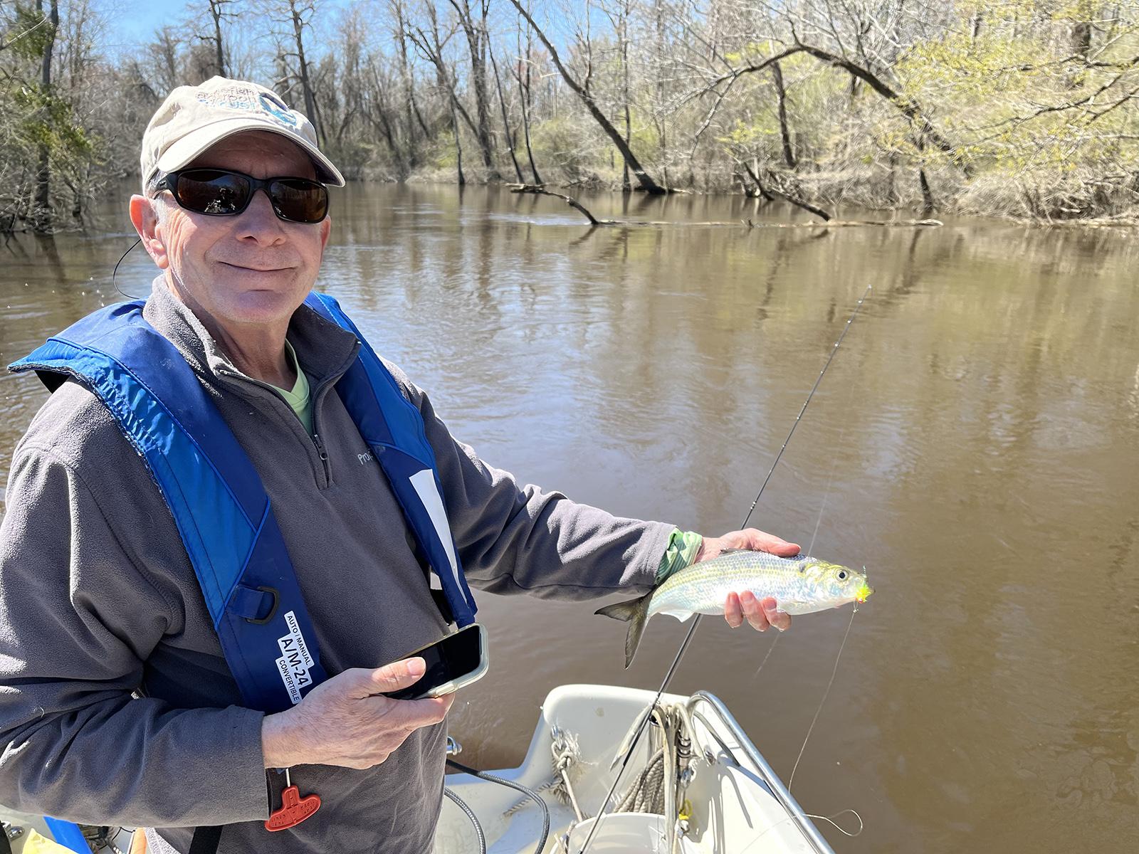 Isang lalaking nakasuot ng life jacket, nakatayo sa isang bangka sa isang ilog at may hawak na isang makintab na shad fish.