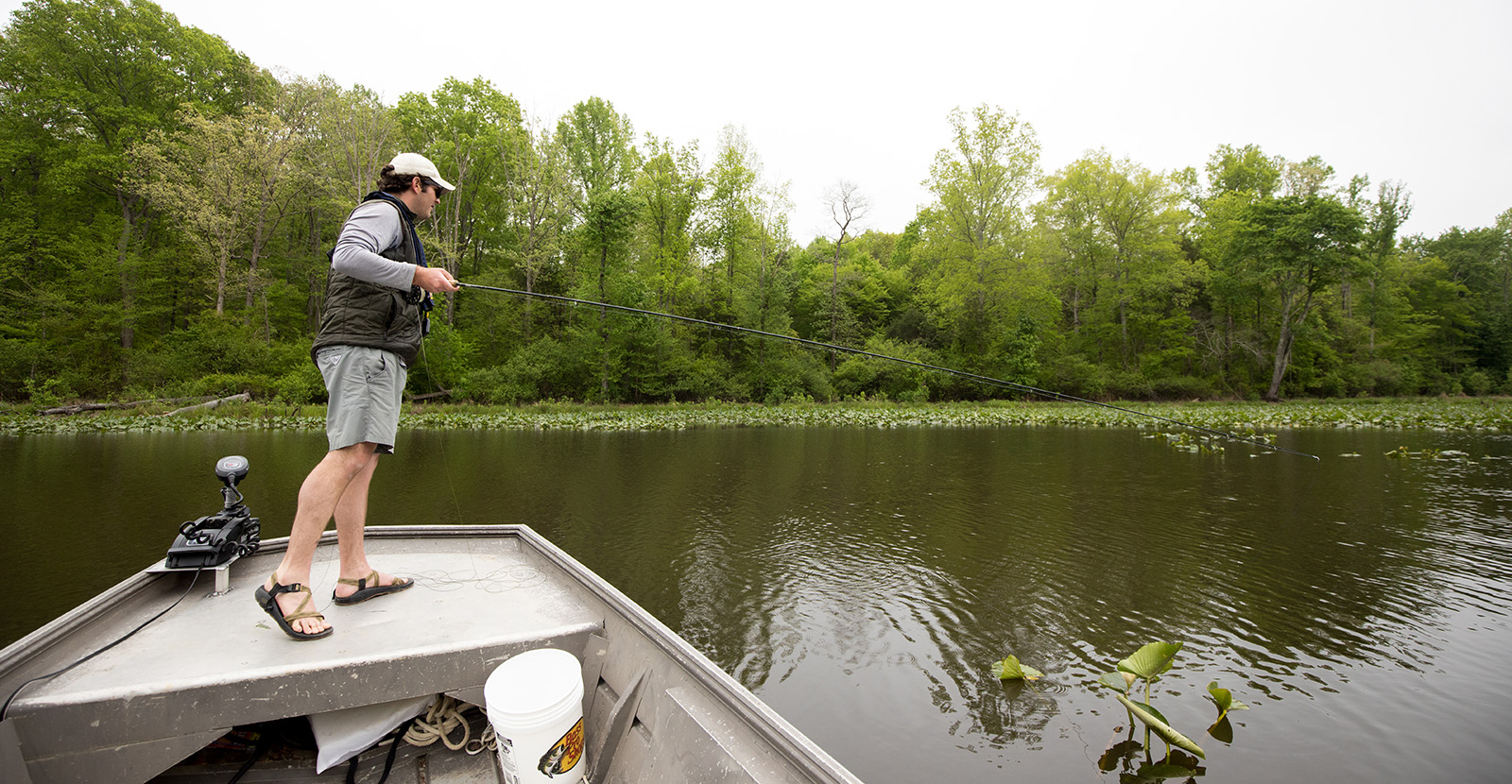 Isang lalaking nakatayo sa busog ng isang johnboat, may hawak na pamingwit at nakatingin sa tubig.