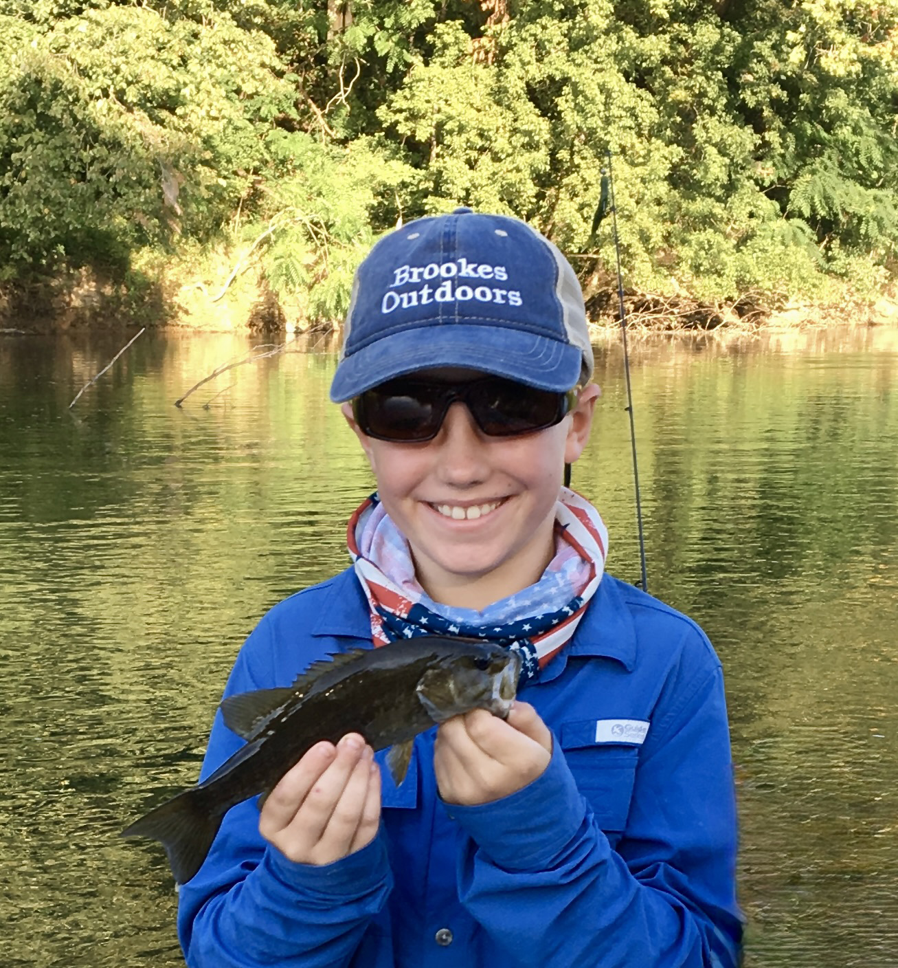 Isang larawan ng isang batang lalaki na may hawak na smallmouth bass at nakangiting may ilog sa likod niya.