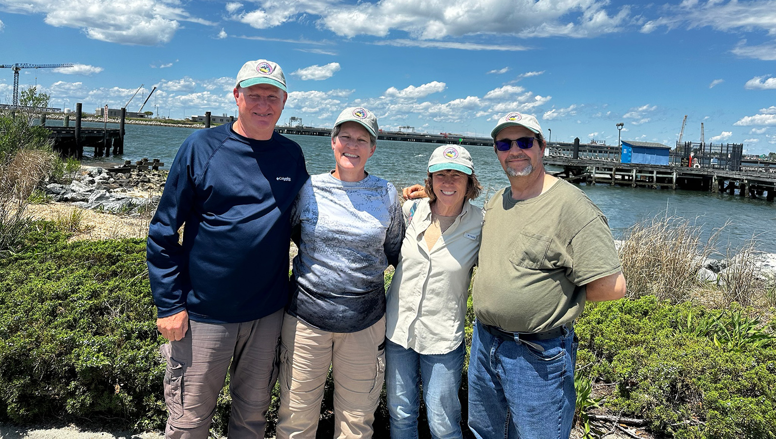 Isang larawan ng apat na nakangiting tao na nakatayo sa dalampasigan ng isang isla na may tubig sa background.