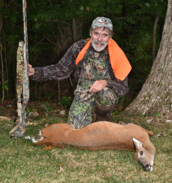 Isang larawan ng isang lalaking naka-camouflage at orange na may hawak na baril at nakaluhod sa likod ng isang whitetailed doe sa lupa.