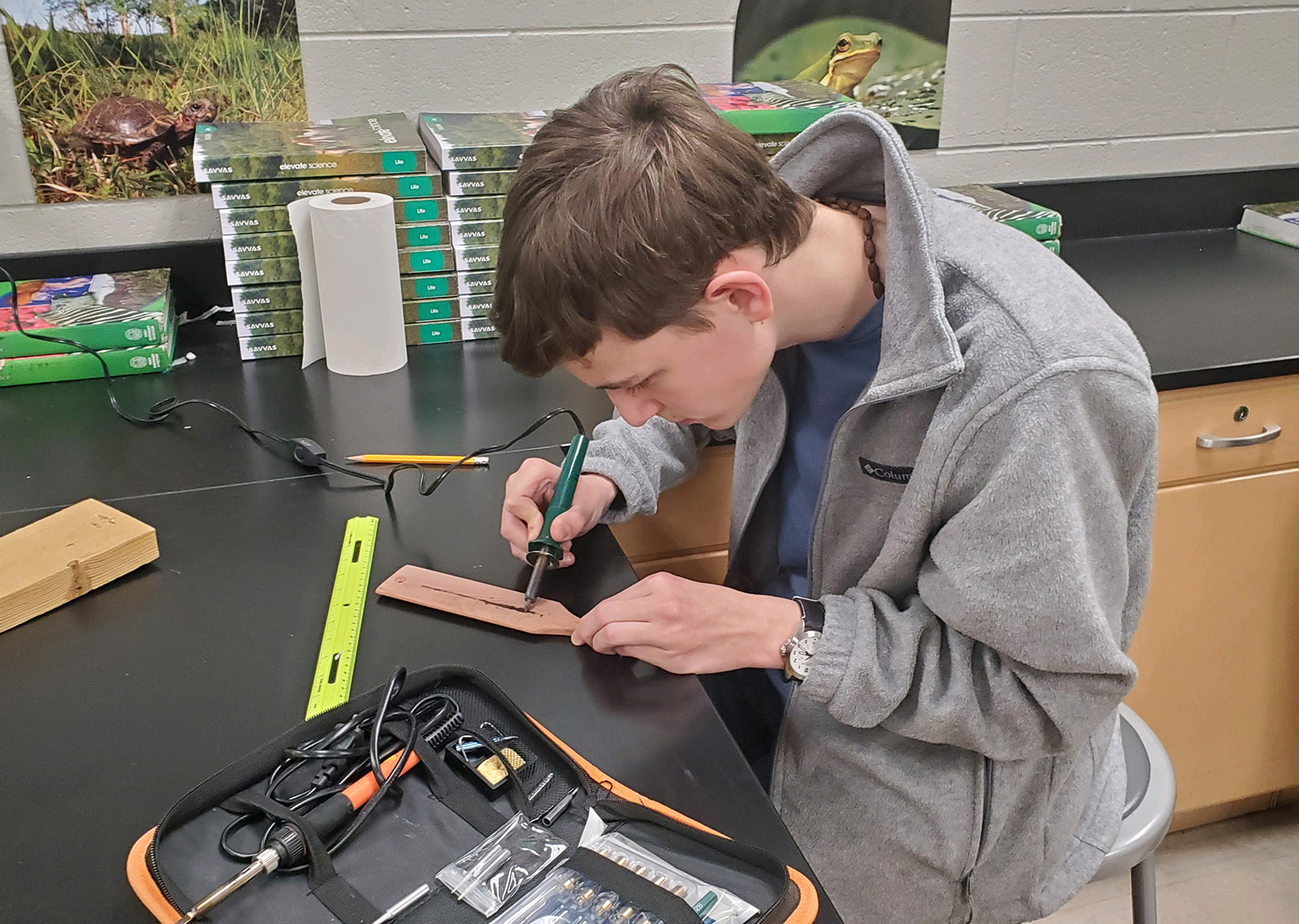 Isang larawan ng isang binata sa isang worktable, gamit ang wood-burning tool sa turkey call paddle.