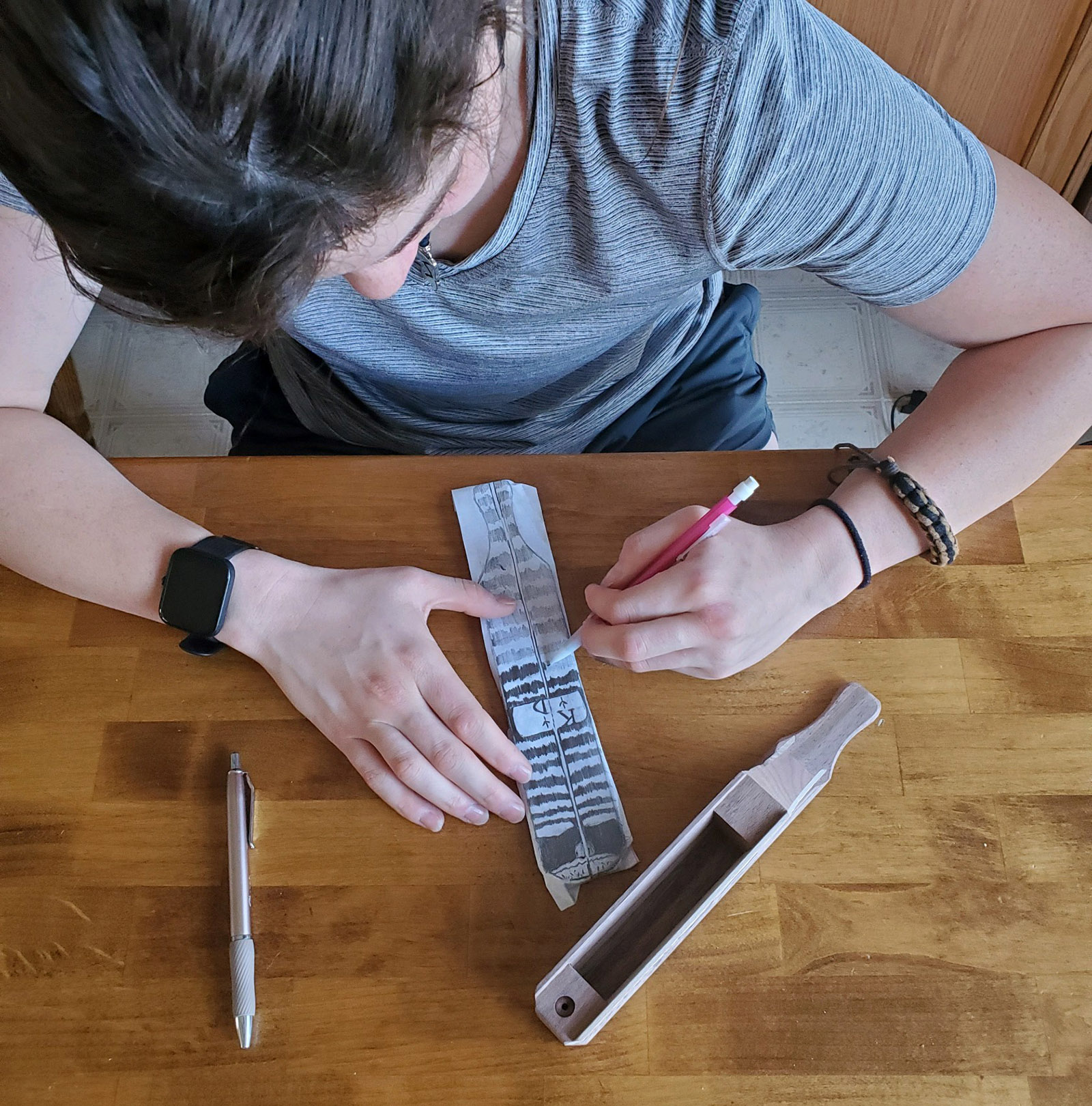 Isang larawan ng isang batang babae na sumusubaybay sa isang pattern sa papel na may paddle turkey call na nakaupo sa tabi niya sa mesa.