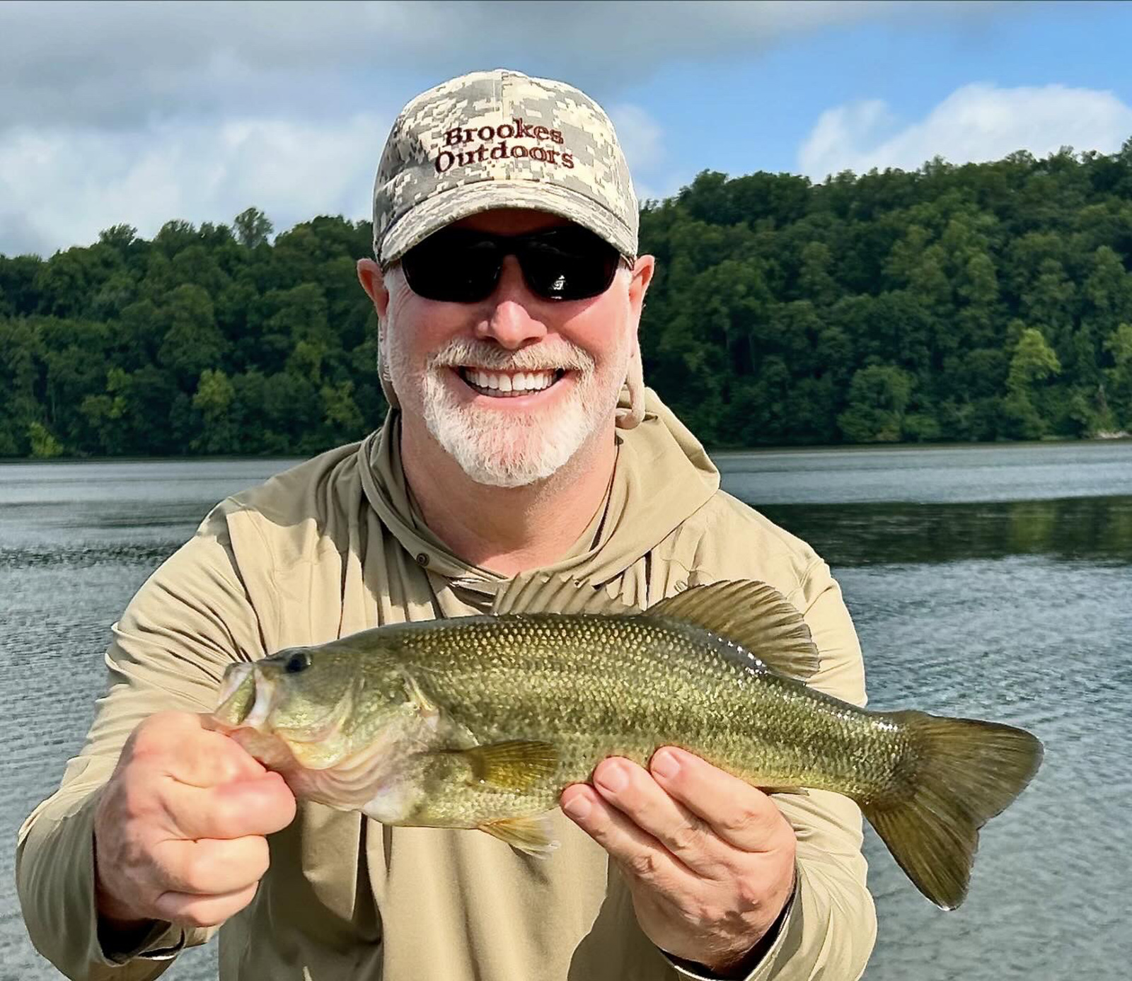 Isang lalaking may hawak na largemouth bass sa harap ng isang ilog.