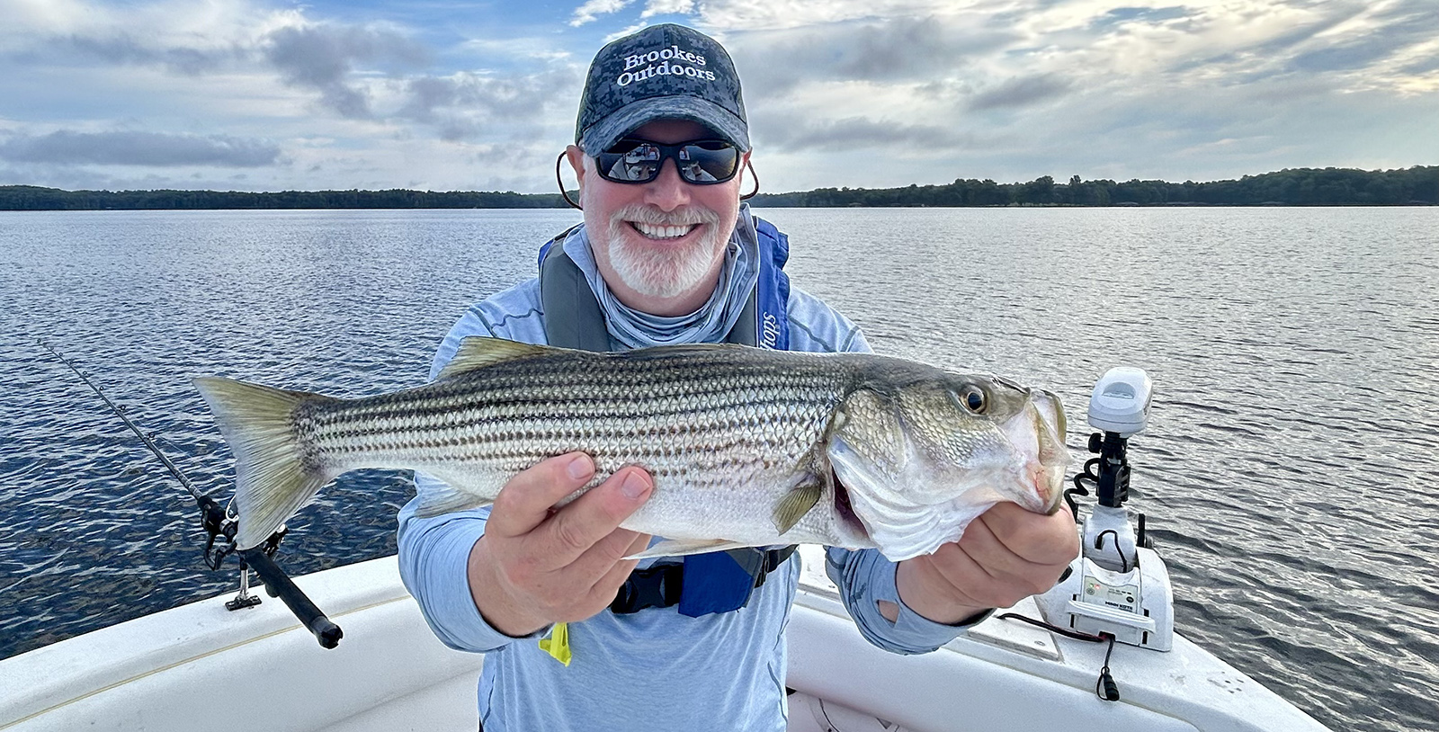 Isang lalaking nakaupo sa isang bangka, may hawak na malaking striped bass at nakangiti.