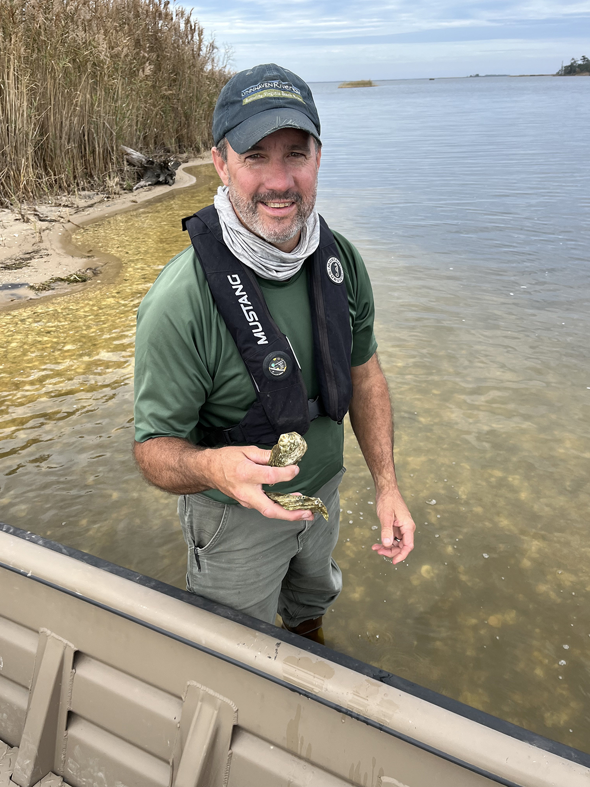 Larawan ng isang lalaking nakatayo sa tubig na hanggang tuhod ang lalim sa tabi ng isang bangka, na may hawak na mga balat ng talaba. 