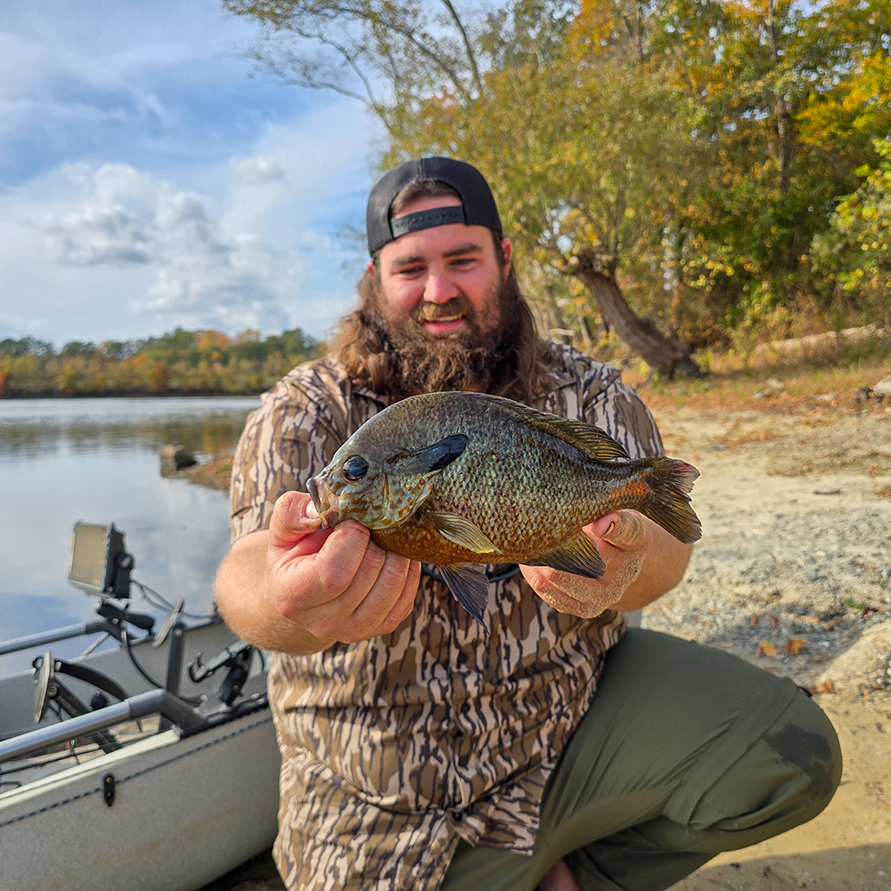 Isang larawan ng isang lalaking nakaluhod sa harap ng isang kayak sa dalampasigan, nakataas ang isang malaking redbreast sunfish at nakangiti.