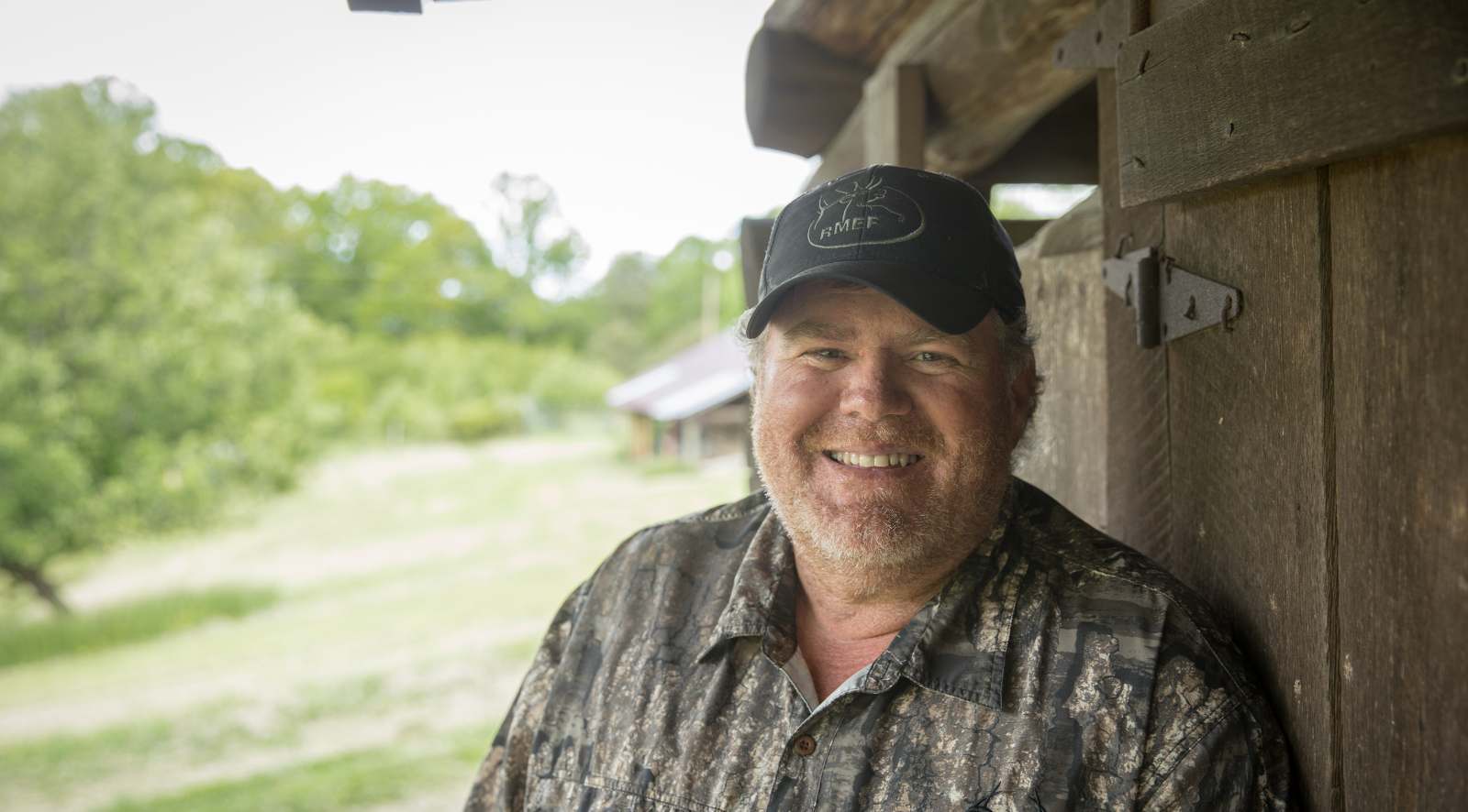 Isang portrait na larawan ng isang lalaking may balbas na nakatayo sa labas sa tabi ng isang magaspang na kahoy na gusali.