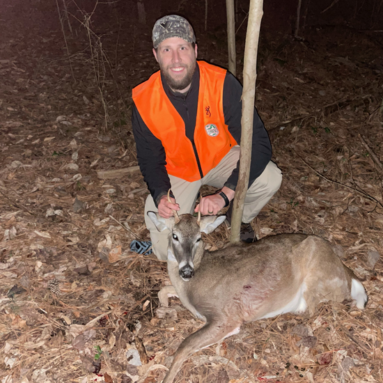 Isang larawan ng isang lalaking nakasuot ng kulay kahel na nakaluhod na may isang spike buck na nakahiga sa lupa sa kanyang harapan,